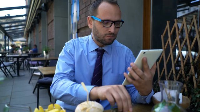 Businessman surfing Internet on tablet and eating hamburger in cafe in city
