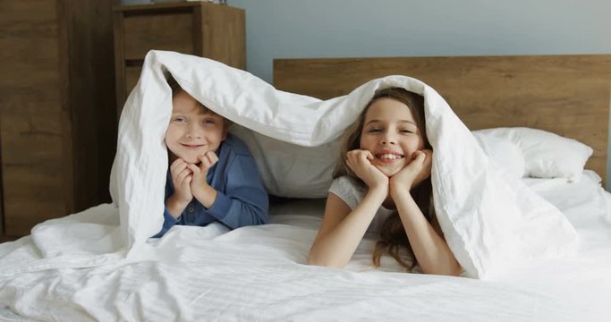 Cute funny little sister and brother son smiling and looking out of the blanket while lying in the bed in the morning. Upside down. Indoor