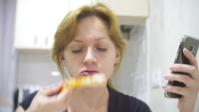 woman is eating pizza in the kitchen at home and using her smartphone, 4k, background blur