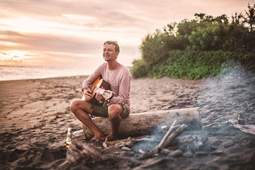 Guy with guitar sitting on log and singing for his friends on summer evening by campfire