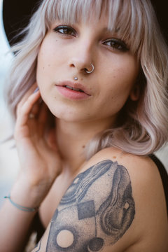 Close up portrait of blonde hipster girl with piercing and tattoos wearing black hat looking at the camera
