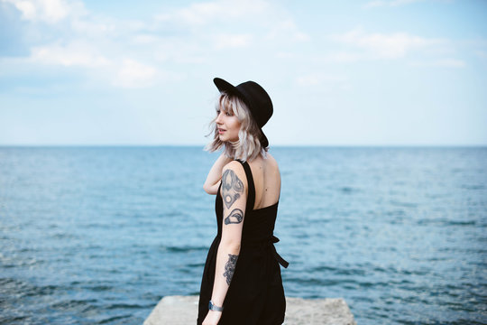 Beautiful blonde hipster girl with tattoos wearing black dress and hat standing on the concrete pier on the sea. Outdoors