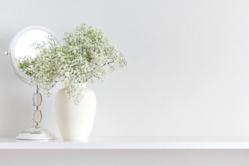 Home interior, floral decor. Beautiful flowers in a vase on a white wall background. White flowers of gypsophila, mirror on a wooden shelf.