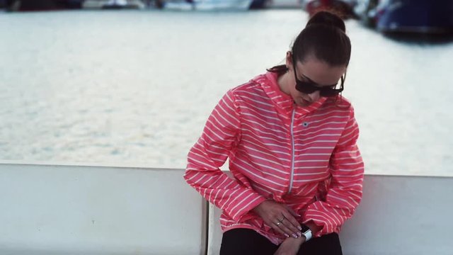 Young girl in orange jacket and sunglasses sitting in sea port and setting up her hand smart watch.