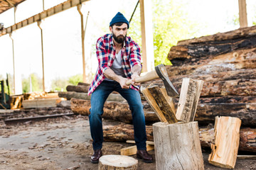 front view of bearded lumberjack in checkered shirt chopping log at sawmill