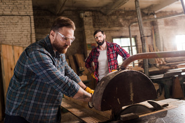 bearded carpenter in safety googles using machine saw and partner with crossed arms standing behind...