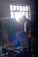 bearded worker in protective gloves wiping forehead and standing near machine at sawmill