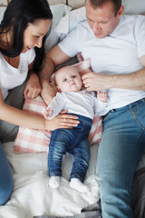 Young mom and dad and their baby son lie in bed and look at the camera indoors. The concept of a happy family