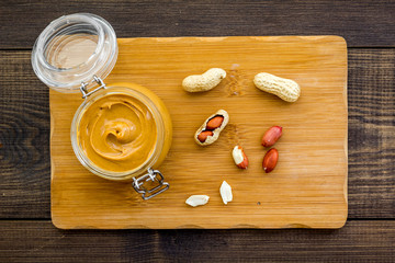 Make peanut butter. Paste in glass jar on cutting desk near peanut on dark woden background top view
