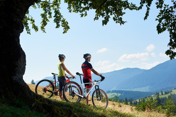Fototapeta na wymiar Active couple sportsman cyclists, man and woman in helmets and full equipment, standing with bikes on grassy hill under big green tree branch, enjoying view of blue mountain and sky on background