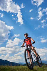 Obraz premium Vertical shot of athletic professional sportsman bicyclist in sportswear and helmet standing with cross country bike, enjoying view of mountains and blue sky with white clouds on sunny summer day