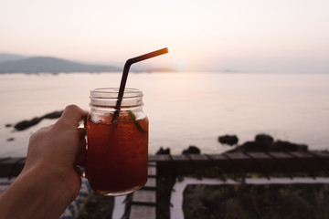 Hand holding glass of lemon ice tea on tropical sea beach in sunset.