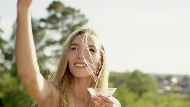 beautiful blonde with long hair smiling, posing and making selfi in brown swimsuit and bikini with wineglass of cocktail a background of nature