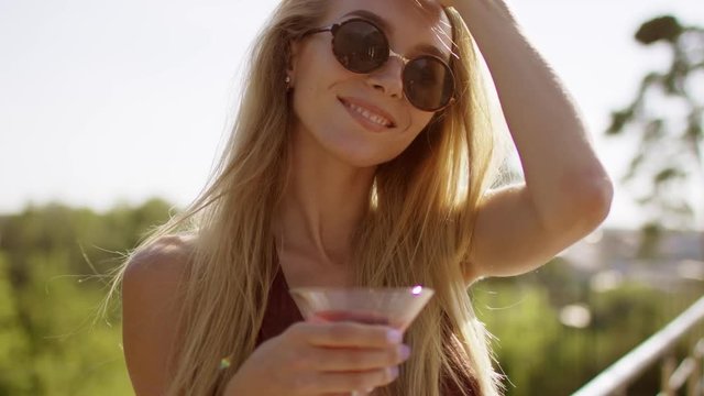 beautiful blonde with long hair smiling, showing off and flirting straightening hair in brown swimsuit and bikini with wineglass of cocktail a background of nature