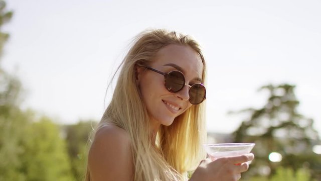 beautiful blonde with long hair smiling, flirting showing off and turning in brown swimsuit and bikini with wineglass of cocktail a background of nature