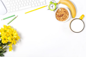 White desktop with keyboard, mouse, colorful stationery, yellow flowers, plate with granola, banana and large cup of milk. Concept of healthy eating at office. Top view.