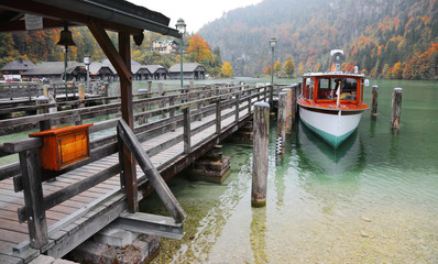 Fototapeta na wymiar Sightseeing boat parking by a wooden pier at beautiful lakeside in a misty foggy morning on Lake Konigssee ~ Beautiful Autumn scenery of Koenigssee (King's lake) in Bavaria, Germany