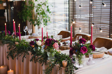 Fototapeta na wymiar Coziness and style. Modern event design. Table setting at wedding reception. Floral compositions with beautiful ranunculus flowers and greenery, candles, laying and plates on decorated table.