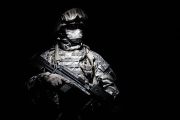 United States Armed Forces soldier in battledress with black glasses and mask on face, armed squad automatic weapon emerges from darkness. Military threat, secret stealth mission, hybrid war combatant