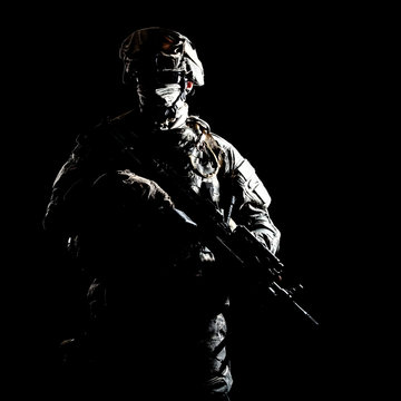US marine raider in combat uniform with hidden face, armed with assault carbine low key, high contract studio shot on black background. Equipped army soldier standing in darkness with weapon in hands