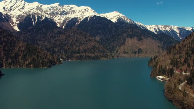 Aerial view of landscape with huge clear lake and mountains. Tops are covered by snow, and slopes are covered pineries, sun is shining bright