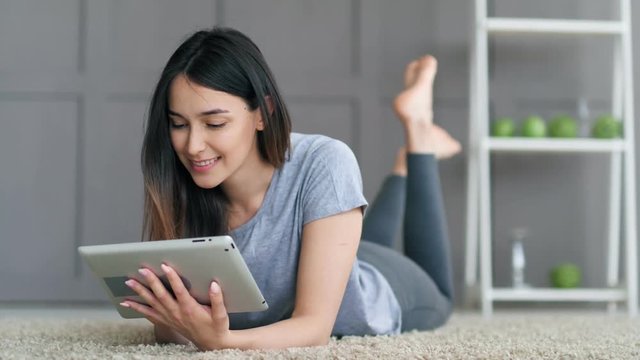 Young happy woman lying on the floor and using tablet computer at home