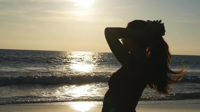 Young brunette woman tying ponytail on beach near the sea at sunset. Beautiful girl squeeze her hair at ocean shore at sunrise. Female tourist braiding hair and making hairdo. Haircare Slow motion