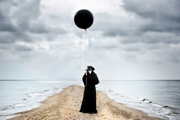 Plague doctor with black balloon in seaside. Outdoor portrait with dramatic sky in background.
