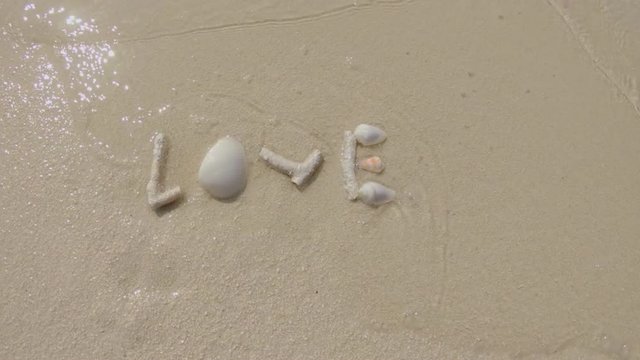 Gentle Sea Waves Wash Over The Word Love Created With Sea Shells And Corals On A White Sandy Beach.