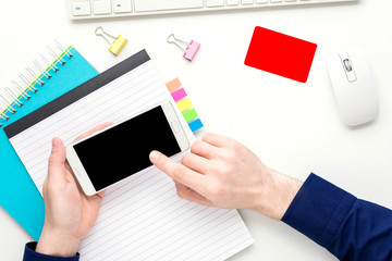 white desk, male hands, guy uses white smart phone, business card lies on the table, office supplies, Notepad, keyboard, mouse, white background with copy space, for advertisement, top view