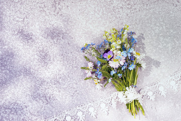 bouquet of wild flowers as forget me not and lily of the valley lays on the silver textured background with lace diagonal frame, top view with copy space