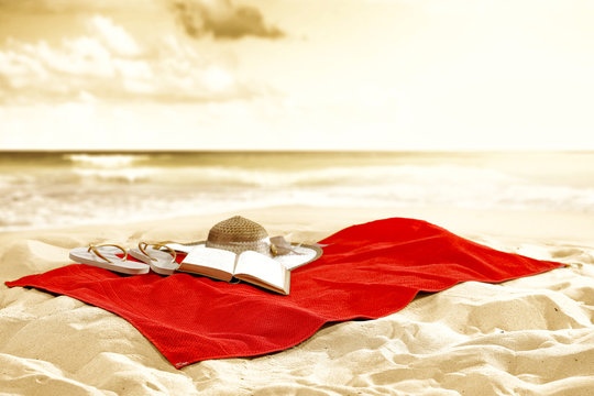 Red towel with an open book, a straw hat abd flip-flops. Summer beach background and copy space for products.