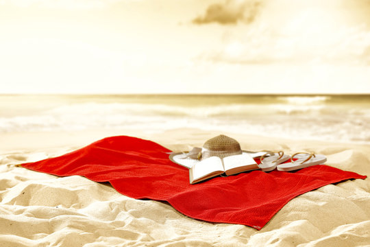 Red towel with an open book, a straw hat abd flip-flops. Summer beach background and copy space for products.