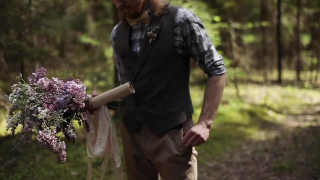 Authentic well-dressed groom holds paper roll letter parcel wild flowers bouquet nature background slow motion. Attractive blonde man festive designer clothing walks in woods forest shallow dof text