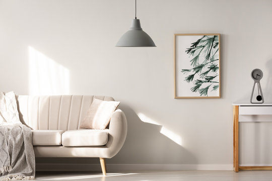 Grey lamp in bright living room interior with poster next to beige sofa. Real photo