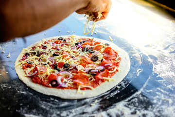 Chef of pizza is garnishing mozzarella cheese on a top or surface of pizza. He is cooking and preparing for baked. The ingredients of pizza have pepperoni, mozzarella cheese, shallots, black olives