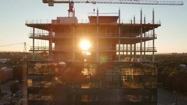 Aerial Shot of the Skyscraper Building in the Process of Construction. In the Background Working Crane, Forest, sea and Sunset. Shot on 4K UHD Camera.