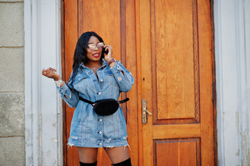 Fototapeta premium African american girl in jeans dress and sunglasses speaking on mobile phone and posed on streets of city. Black stylish model shoot.