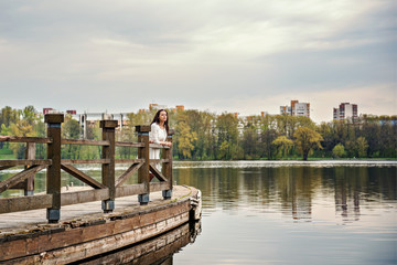Fototapeta premium thinking woman in white dress is standing on a wooden pontoon bridge on a river lake with a cityscape on a background 