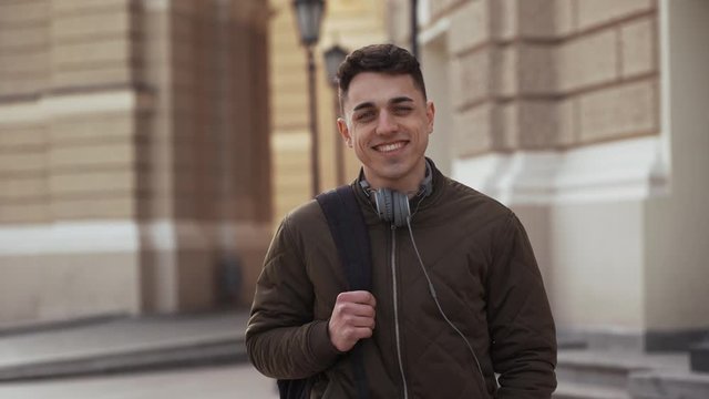 Portrait of caucasian young man 20s wearing jacket walking with backpack after study and posing over old historic building with smile