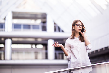 Fototapeta premium Portrait of smiling beautiful young business woman talking on mobile phone, holding on to railing and standing in office hall
