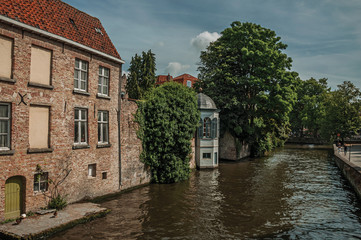 Fototapeta premium Trees and old brick buildings on the canal's edge in a sunny day at Bruges. With many canals and old buildings, this graceful town is a World Heritage Site of Unesco. Northwestern Belgium.
