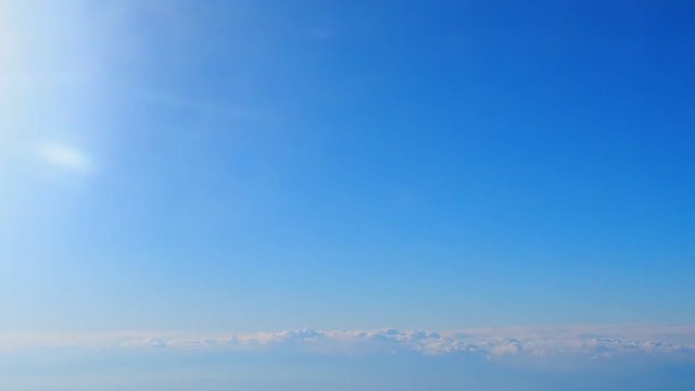 Bright blue clear sky background with small skyline and grey cloud cluster line below moving to the right as view from flying airplane, with white sun shining flare from the left