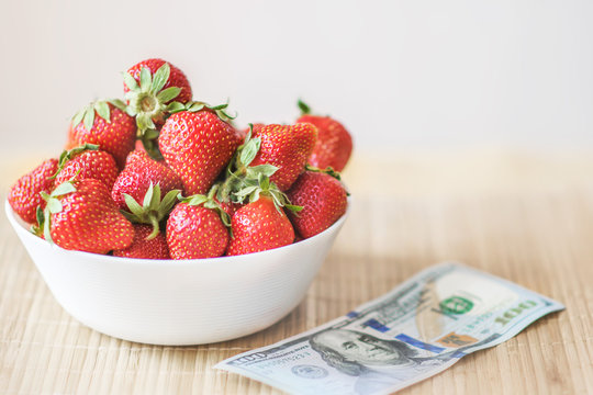 Fresh summer strawberries in a white dish on table with monye banknote one hundred dollars. The concept of healthy food and prices increase