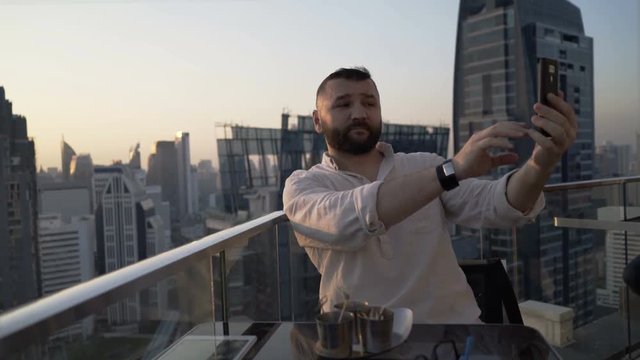 Man sitting on rooftop with the view at the city and doing photos on smartphone
