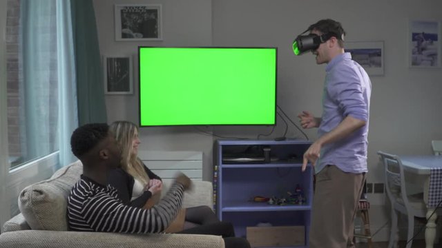 Man on VR headset in Living room friends greenscreen. Handheld shot of millennial group of friends on sofa beside green screen TV, one using VR headset and freaking out