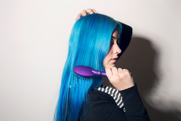 Young woman brushing her long blue hair. Indoors, over a white wall.