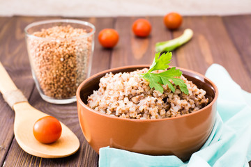 Cooked buckwheat in a bowl with a sheet of parsley, a glass of raw buckwheat and cherry tomatoes on a wooden table