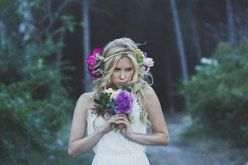 Obraz na płótnie Canvas Upper body portrait of blond Caucasian woman with wavy hair, posing in romantic style in the forest holding flower bouquet. Young girl wearing elegant white dress, flower crown with green background