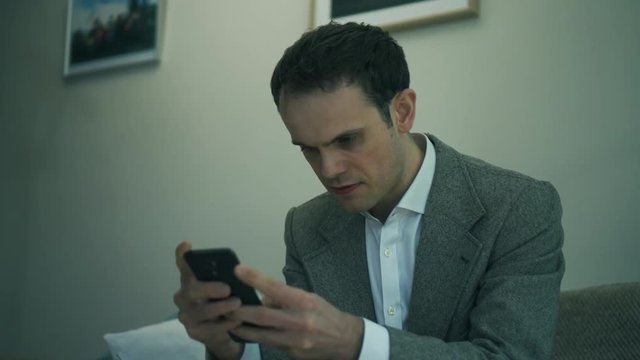 Business man at home furiously texts from smartphone. Handheld shot of business man furiously type on phone from sofa
